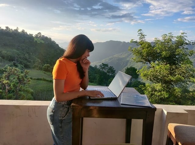 A woman stands on a balcony, working at a desk on her laptop, green hillsides and trees beside her.