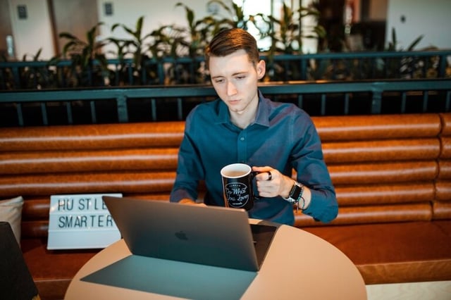 A young professional works from a laptop in a coffee shop, a mug of coffee in one hand.
