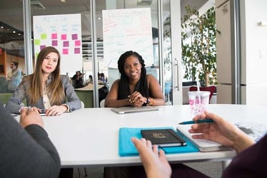 Two young women smile during a meeting as they communicate with their meeting leader.