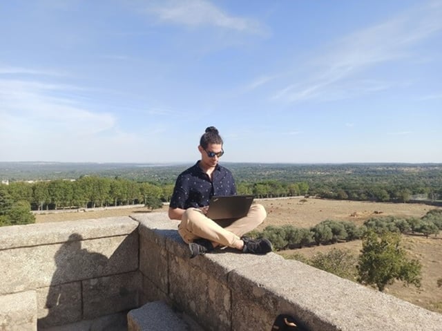 A man sits on a wall, working on a laptop.