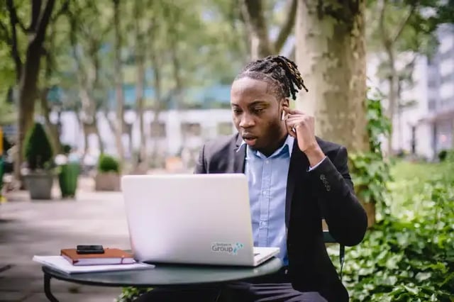 A man sits on a cafe table outside, working on a laptop. The laptop has a The Intern Group logo sticker on it.