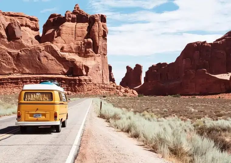 A campervan drives down a road, passing rocks, mountains, and grassy areas as they travel abroad.