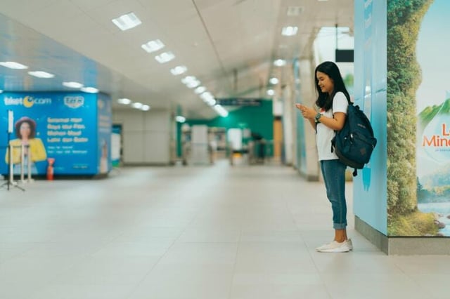 A young woman stands alone in an airport or train station, looking at her phone with a rucksack on her shoulder.