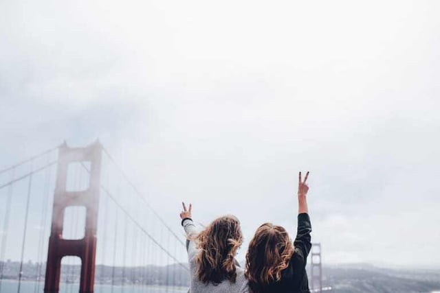 Two women face away from the camera, looking at the mist over the Golden Gate Bridge. The women are holding up peace signs with their fingers.