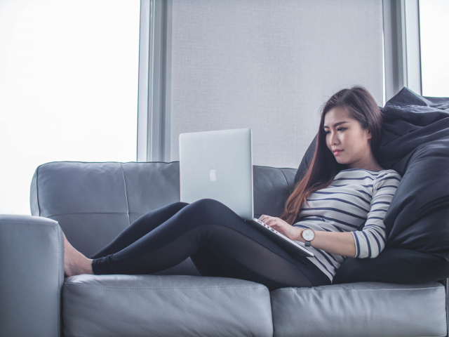 A young woman relaxing on a sofa types on a laptop