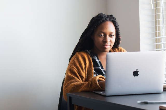 A woman in a blouse and cardigan sits at a laptop with a blank wall behind her