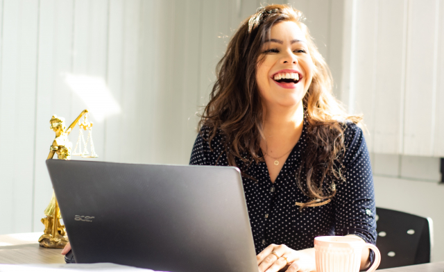A young woman working at a laptop laughs at the camera