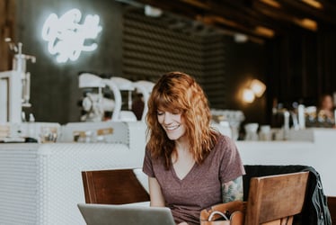 A young professional woman working remotely from a coffee shop. She is smiling at her laptop.