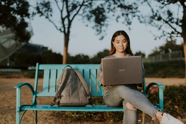 a girl sitting on a blue bench with a backpack next to her, working on a laptop