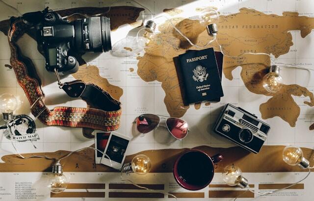 A world map laid out on a table. On top is a camera, a passport, a cup of coffee, and fairylights.