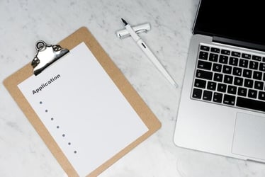 A picture of a desk with a laptop and an application form with a clipboard.