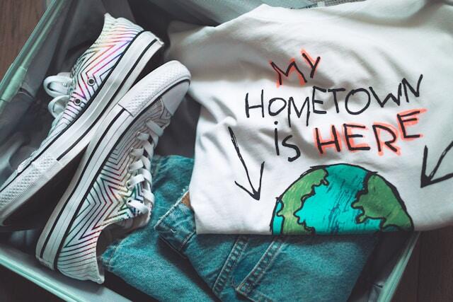 An open suitcase with clothing and shoes. A t-shirt says 'my hometown is here', pointing to a globe.