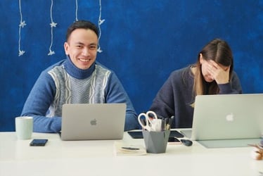 Two professionals work together on laptops. One of them looks confident at work, and the other looks upset.