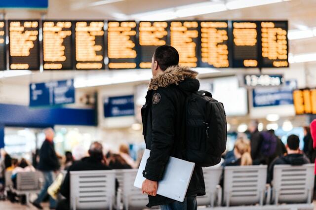 A man looks at a departure board in a train station. He is wearing a coat and a rucksack and has a laptop under his arm.