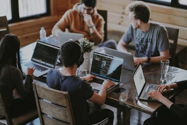 A group of young interns work together on laptops around a table in a coworking space.