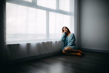 A woman sits in an empty room by a window looking sad.