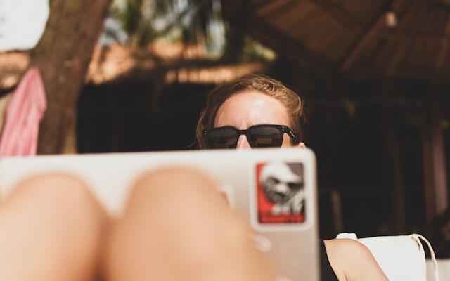 A professional woman works from a laptop whilst sitting on a beach. Her laptop is perched on her lap whilst she lays on a sunlounger and she is wearing sunglasses.