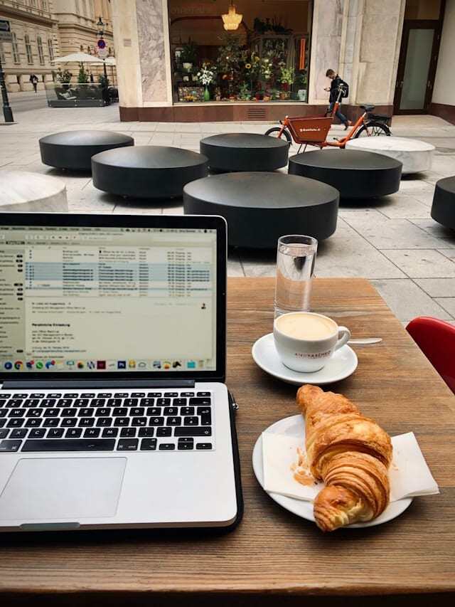 A laptop on a table in a cafe displaying an email inbox on the screen. Beside the laptop is a coffee and a croissant.