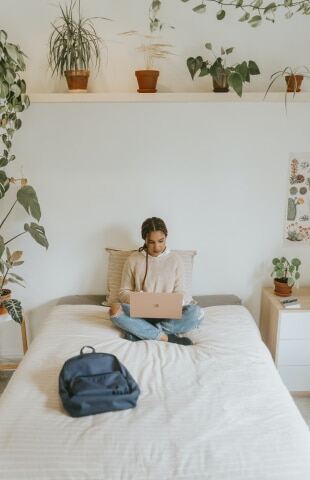 A professional young woman working from home. She is sitting cross-legged on a bed with a laptop on her lap.