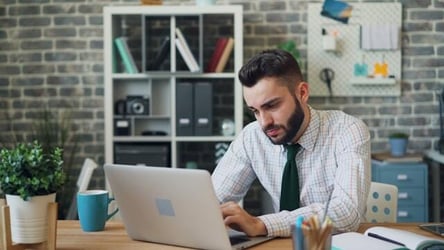 A young professional man in a shirt and tie looks sad whilst working at a laptop.