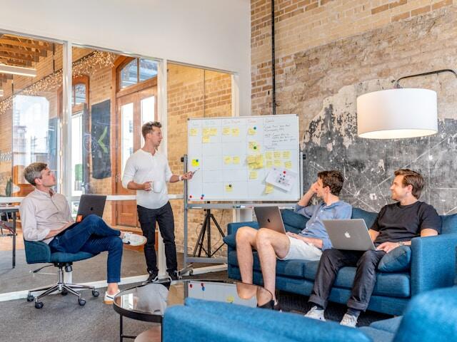 A man gives an elevator pitch to three colleagues in front of a whiteboard full of notes.