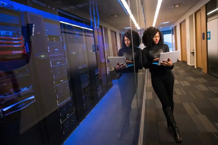 A professional woman stands against a glass wall with a laptop, computer servers in the room beside her.