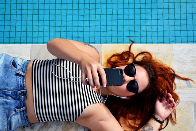 A young woman lies down beside a pool. She is wearing jeans and a t-shirt and is scrolling through her phone.