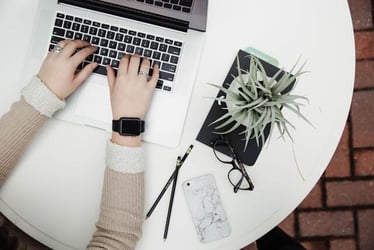A top view of hands typing at a laptop. Beside the laptop is a phone, glasses, pencils, and a potted plant.