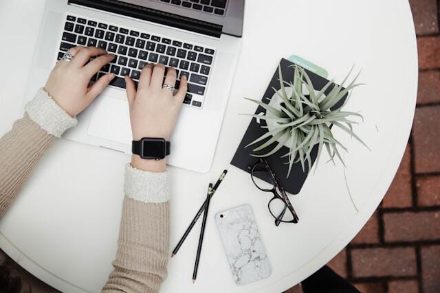 A top view of hands typing at a laptop. Beside the laptop is a phone, glasses, pencils, and a potted plant.