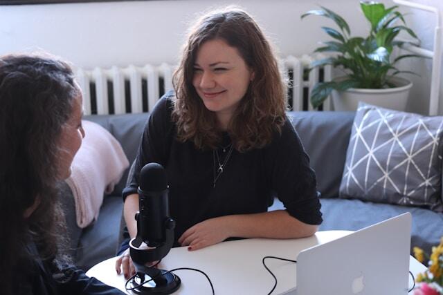 Two women record a podcast or radio show with a microphone and a laptop on a desk.