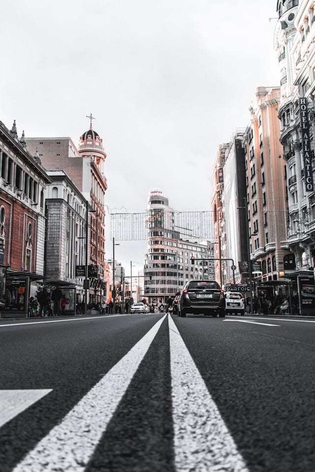 A close up view of a road in Madrid. Cars are driving past in the foreground and there is beautiful Spanish architecture in the background