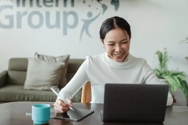 A smiling woman works at a desk, laptop and tablet in front of her. The back wall is printed with The Interrn Group logo.