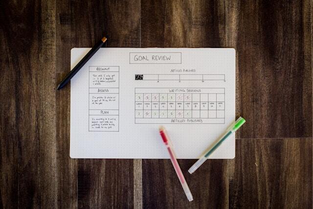 A top down view of a notebook on a table. The notebook features a neat work tracker and three pens on top.