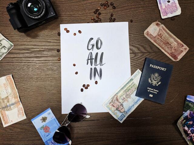 A piece of paper on a table which reads 'Go all in.' Around the paper is foreign currency, a passport, sunglasses, and a camera