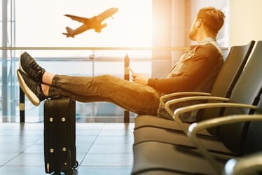 A young man sits in an airport with his feet on his luggage. He looks towards the window where a plane is taking off.