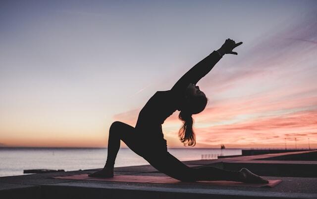 A woman does some yoga outside overlooking a lake, the sun rising behind her.