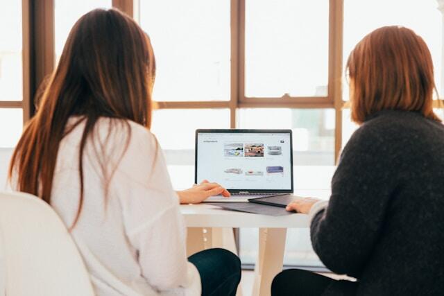 Two women face away from the camera, asking questions and working on a laptop and tablet.