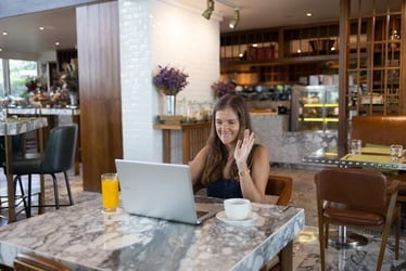 A woman introduces herself on an online learning course. She waves whilst sitting at a table with a laptop.