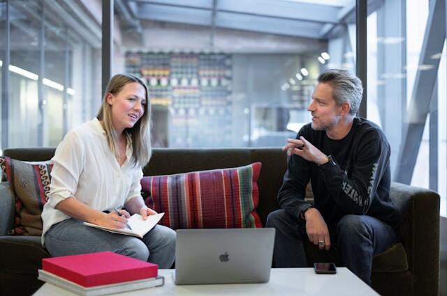 Two professionals sit in a relaxed office, talking and working from laptops and notebooks.