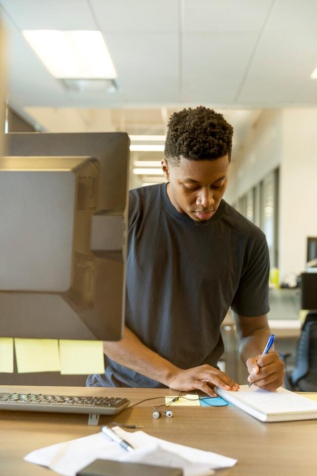 A man stands working at a desk. There are sticky notes on his computer and he's writing in a notepad.