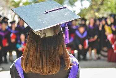 A graduate standing facing away from the camera to address a crowd. She is wearing graduation robes and a mortarboard.