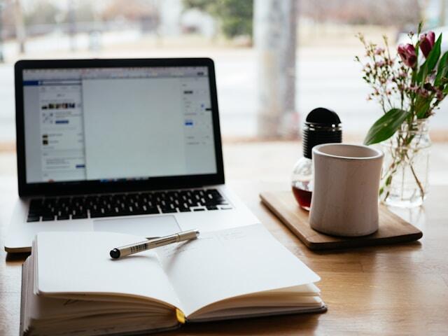 A laptop on a desk displaying a social media account. In front of the laptop is an open notebook and pen.