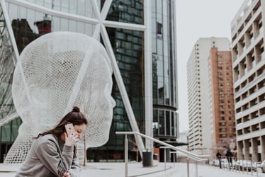 A young woman sits outside, speaking on the phone somewhere abroad. There is a large, modern artwork beside her in the shape of a head and face.