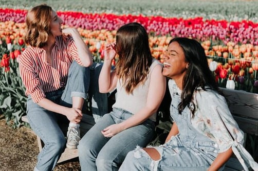 Three young professional women laugh together on a bench in front of a tulip field.