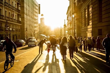 People walk through a street in Madrid, Spain, the sun setting or rising through the buildings to bathe everything in a warm light.