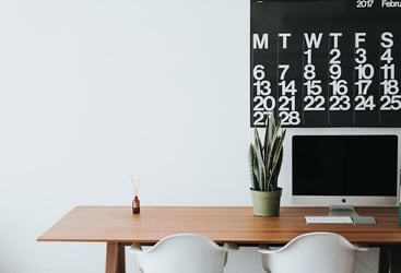 A desk with a computer and a calendar behind it.