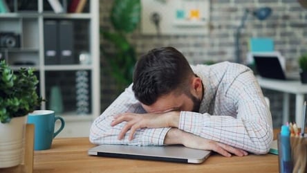 A man with a laptop at a desk rests in head on his arms in stress over his laptop.