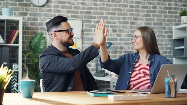 Two coworkers high five each other at their desk.